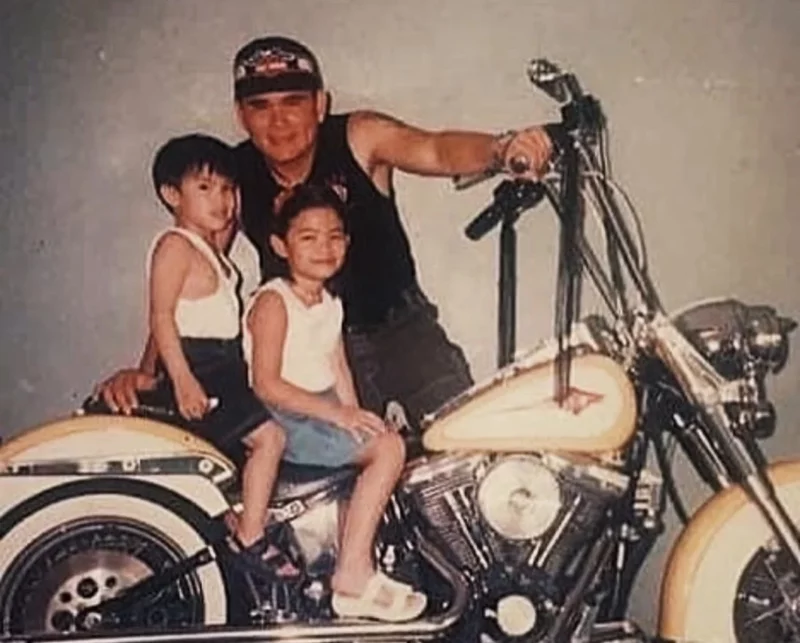 A nostalgic, vintage childhood photo of a young Katrice Kierulf and her brother sitting on the seat of a classic cream-and-black cruiser motorcycle, while their father, Daddy Lee, stands behind them in a black tank top and cap, smiling and holding the handlebars.